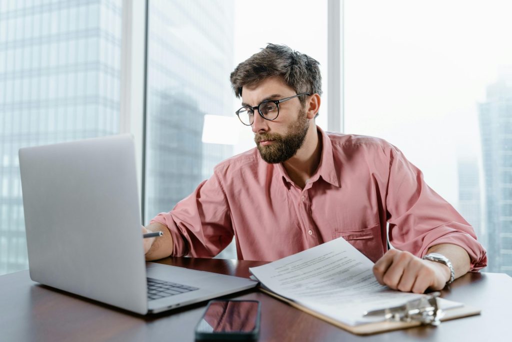 Ein Geschäftsmann überprüft in einem Büro Dokumente und Daten auf einem Laptop und konzentriert sich auf die Finanzanalyse.