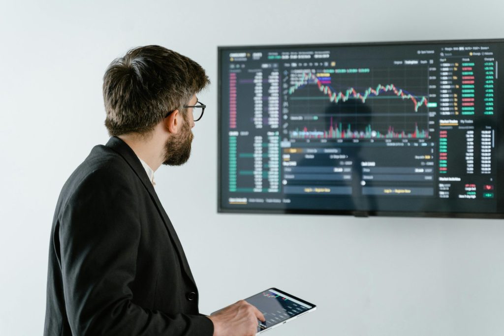 Mann beobachtet Börsendaten auf einem Bildschirm und hält ein Tablet, das Finanzanalysen und -handel darstellt.