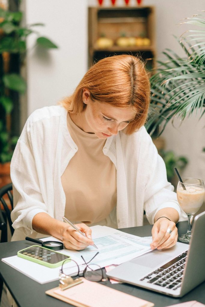 Frau analysiert Finanzdokumente mit Laptop und Taschenrechner im Haus.